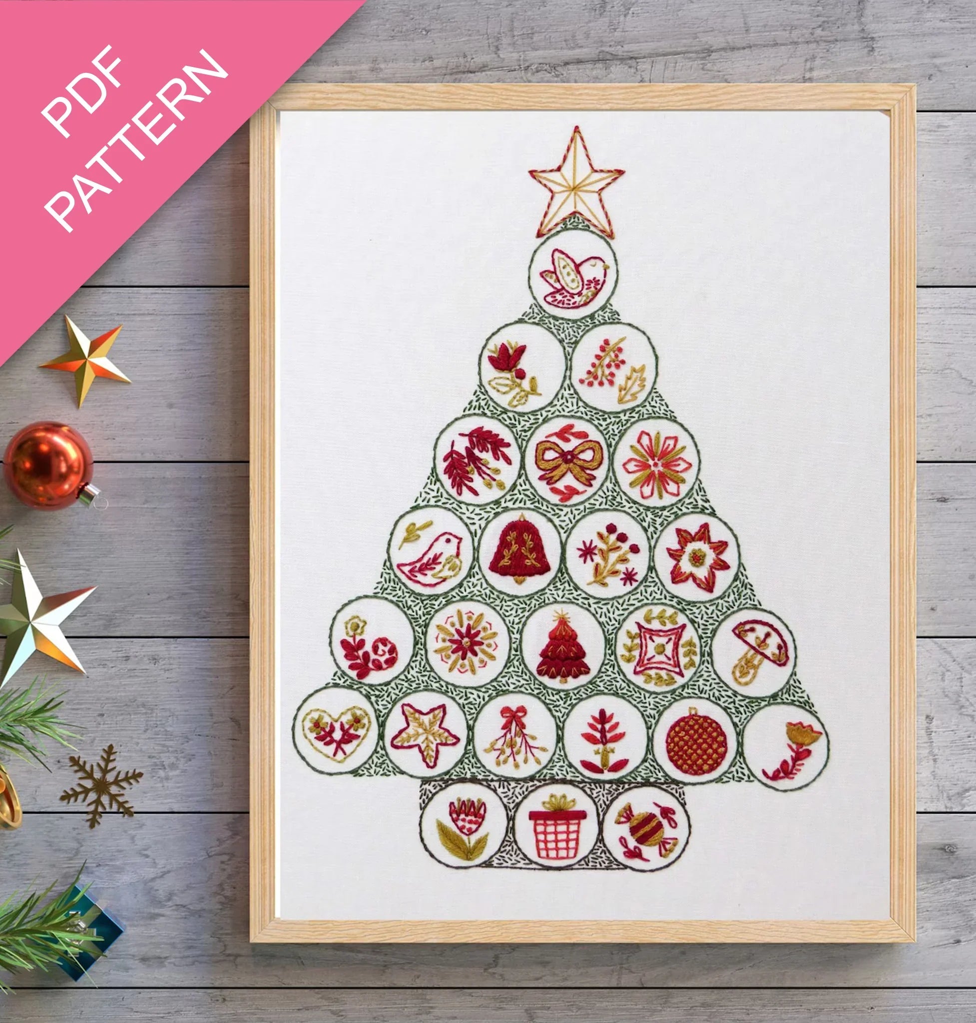 Framed cross-stitch pattern in the shape of a Christmas tree on a wooden surface with decorative items.
