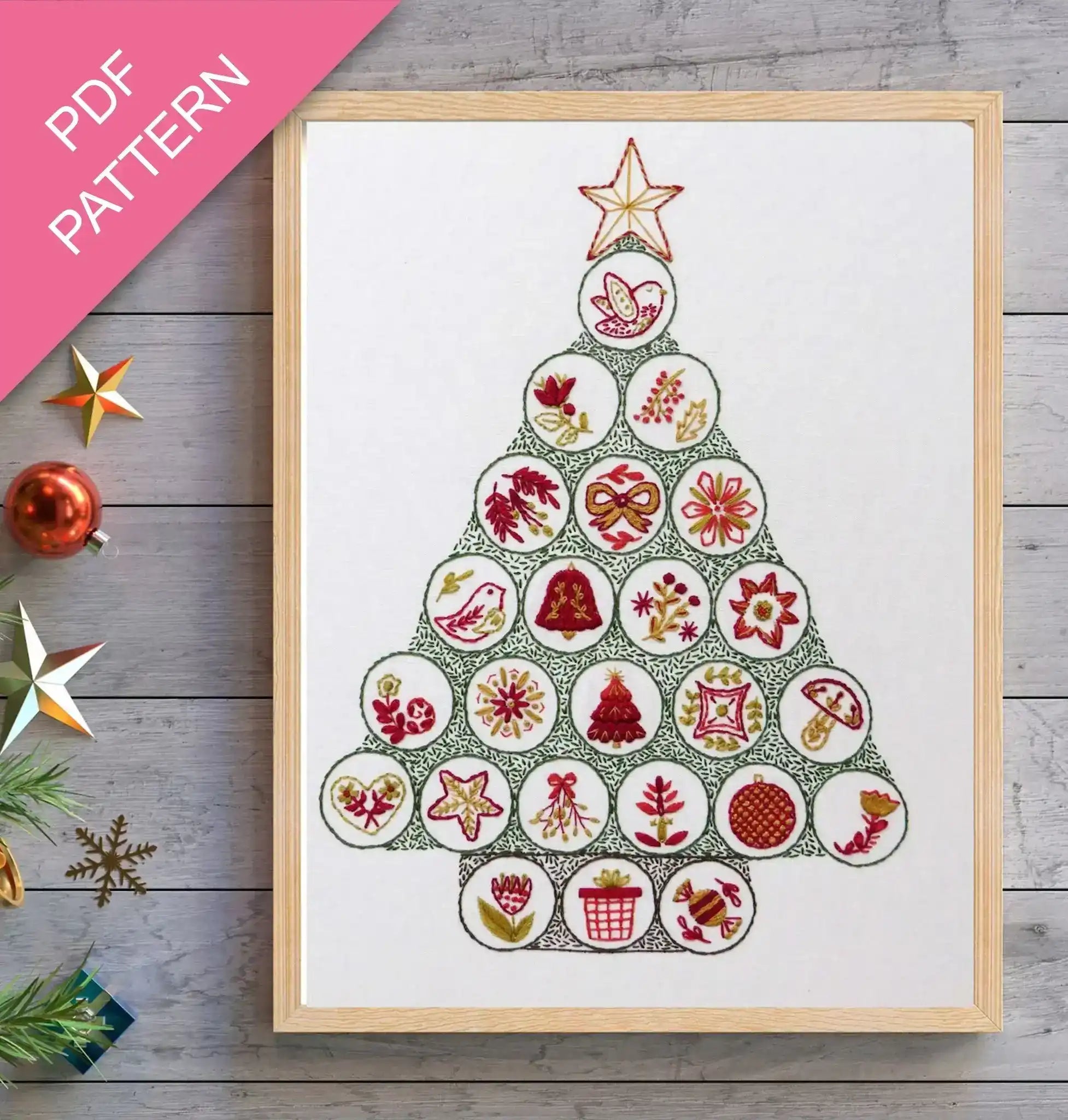 Framed cross-stitch pattern in the shape of a Christmas tree on a wooden surface with decorative items.