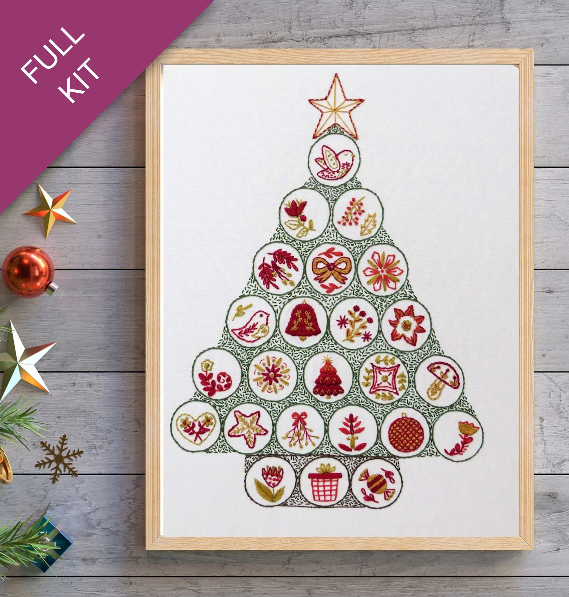 Framed cross-stitch pattern of a Christmas tree on a wooden surface with decorative items.