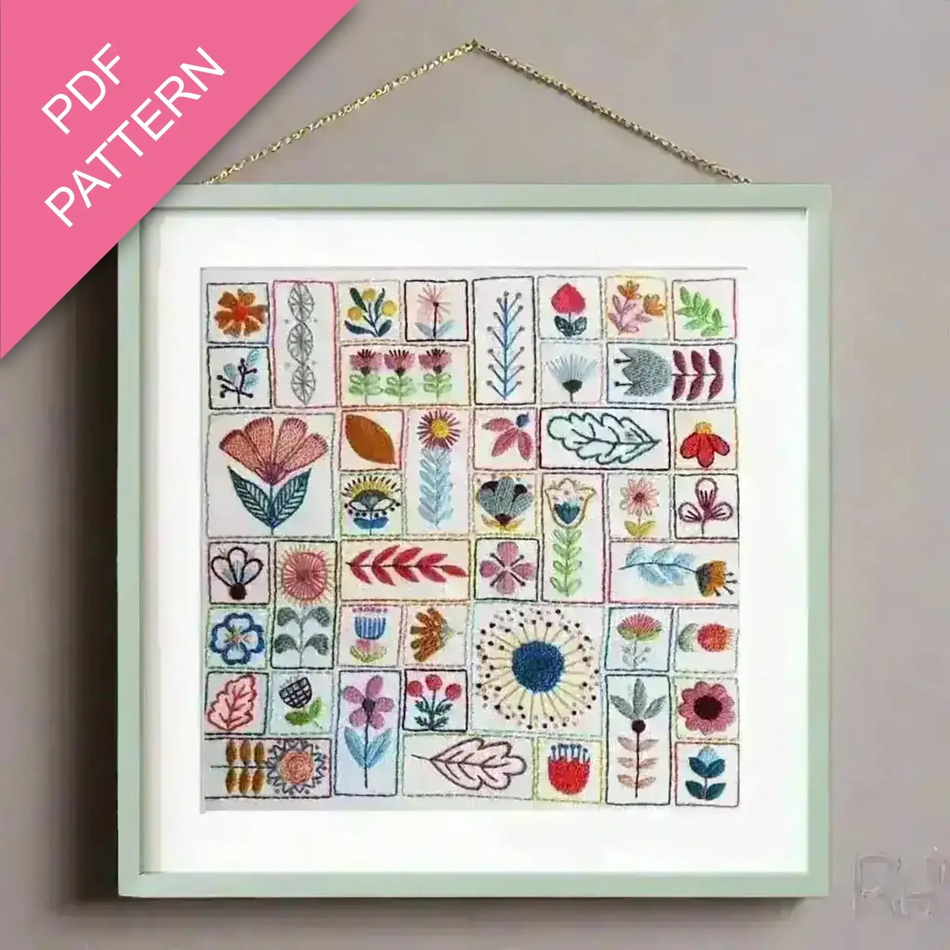 Framed embroidery pattern with floral designs on a plain wall, featuring a pink PDF pattern overlay.
