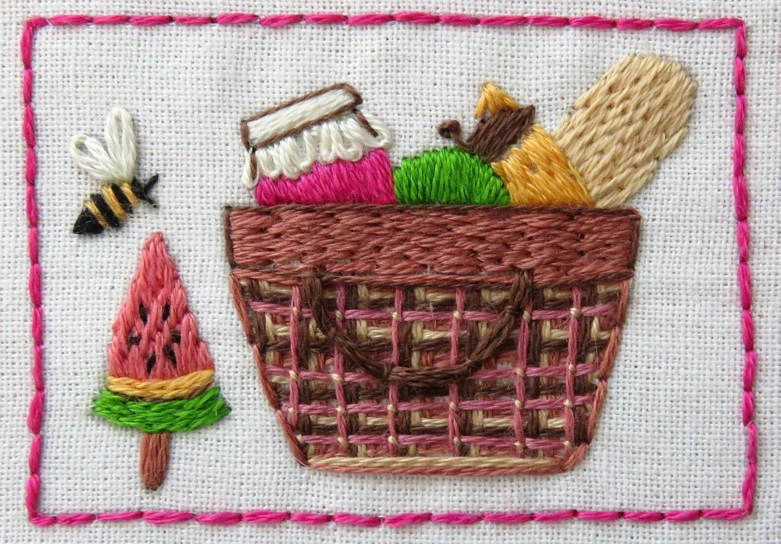 Embroidered design of a picnic basket with food items and a watermelon, framed with pink stitching.