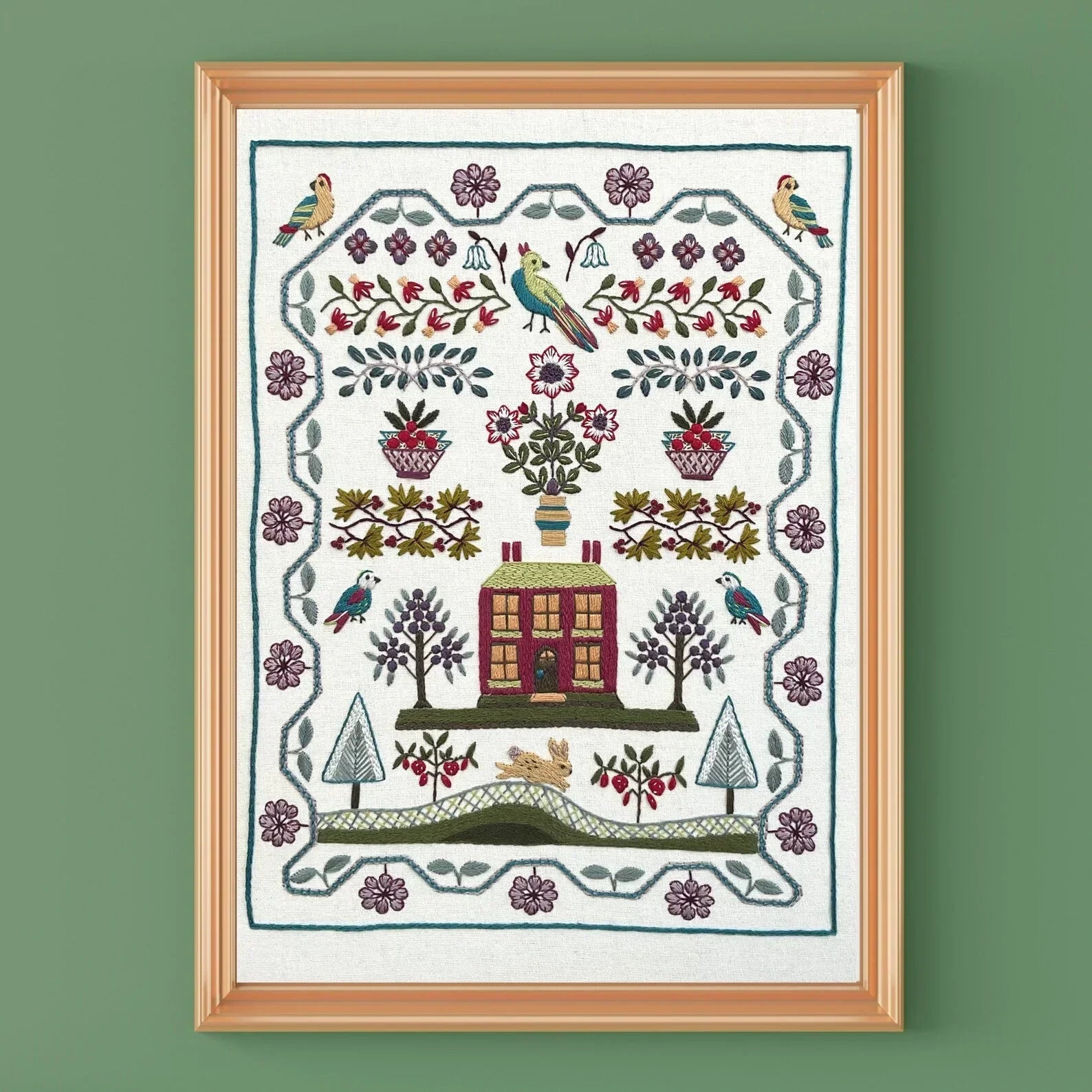 Framed embroidered artwork with a house, trees, and birds on a green wall