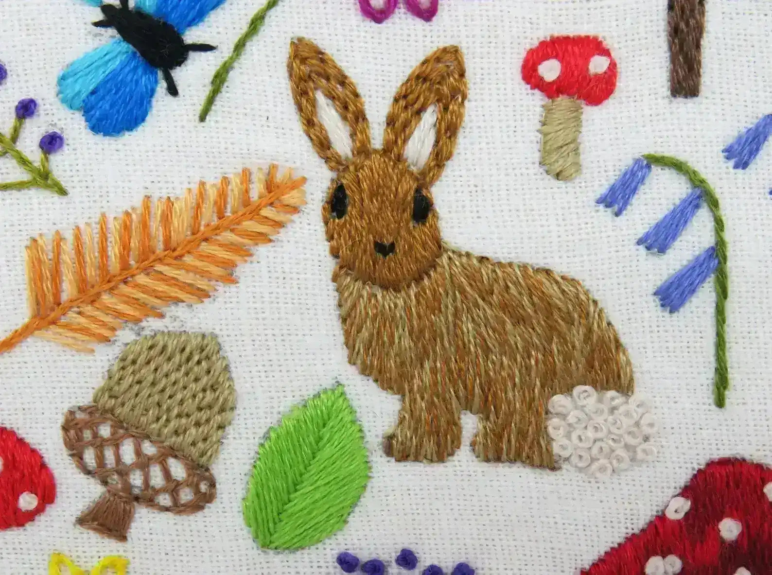 Embroidered design of a rabbit surrounded by flowers, leaves, and mushrooms on a white background.