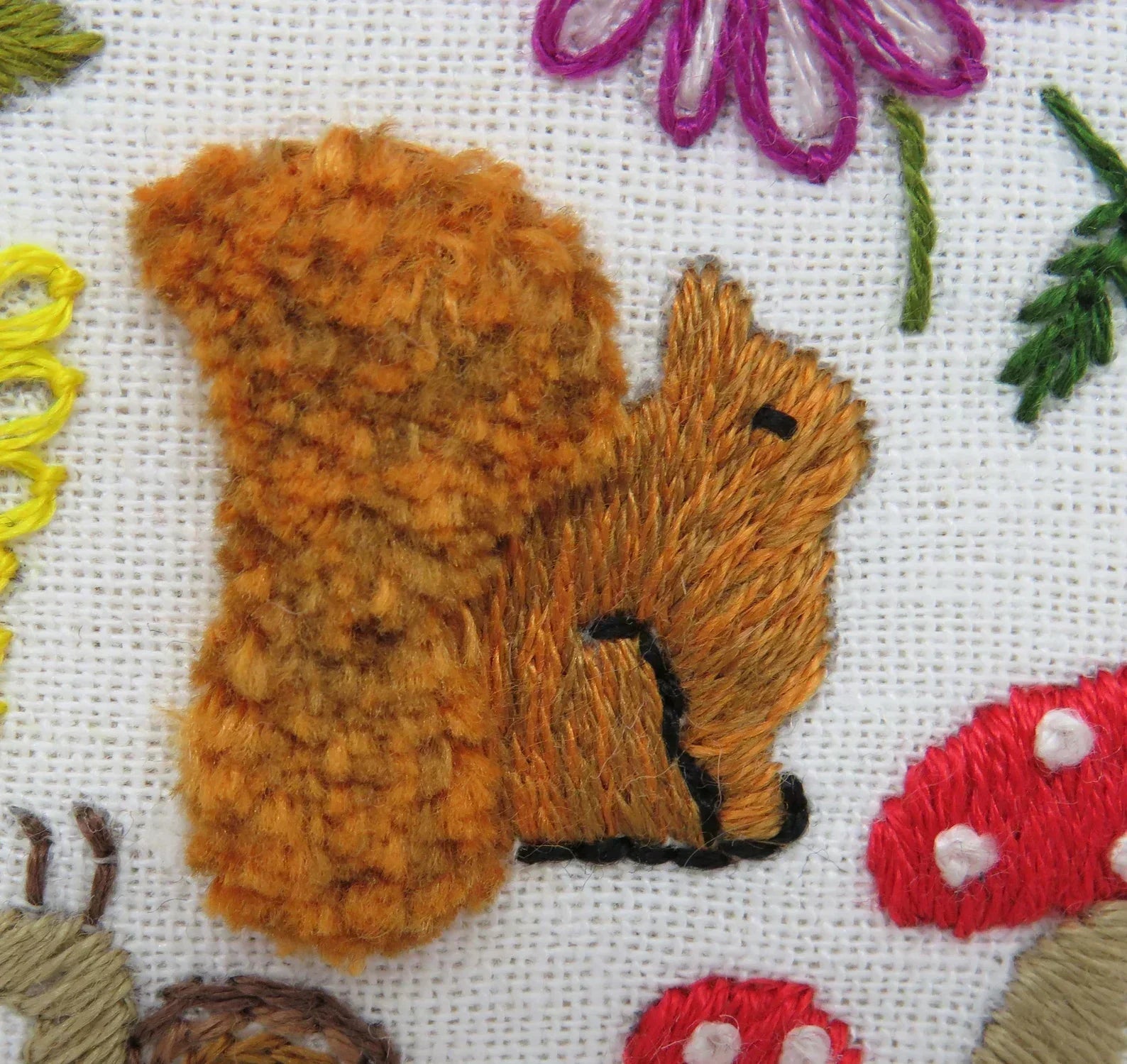 Embroidered design of a brown squirrel surrounded by flowers and mushrooms on fabric.