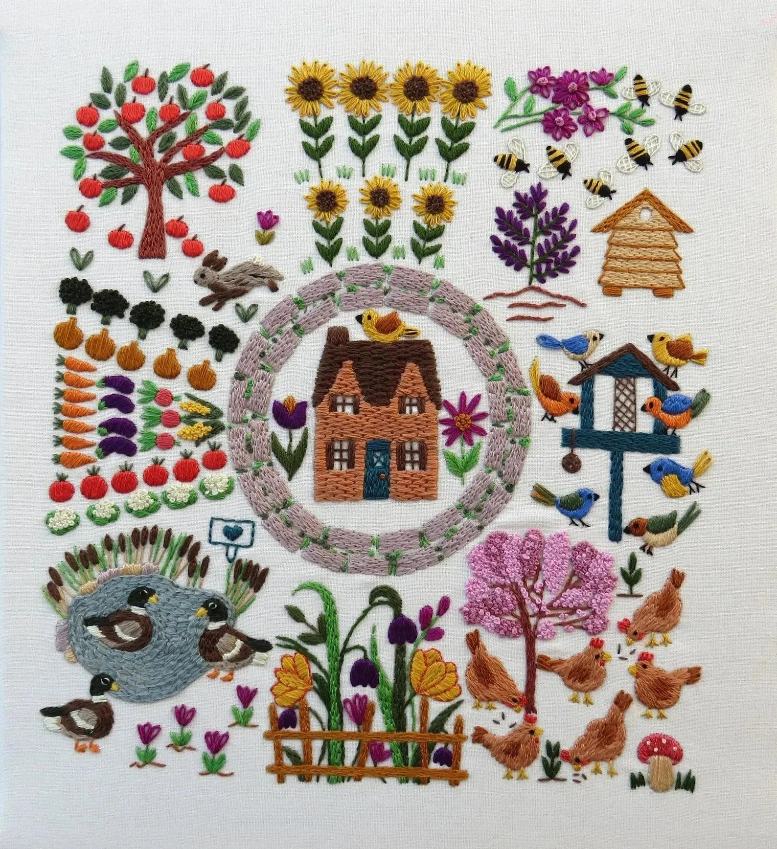 Embroidered design of a garden scene with house, animals, and plants on a white background