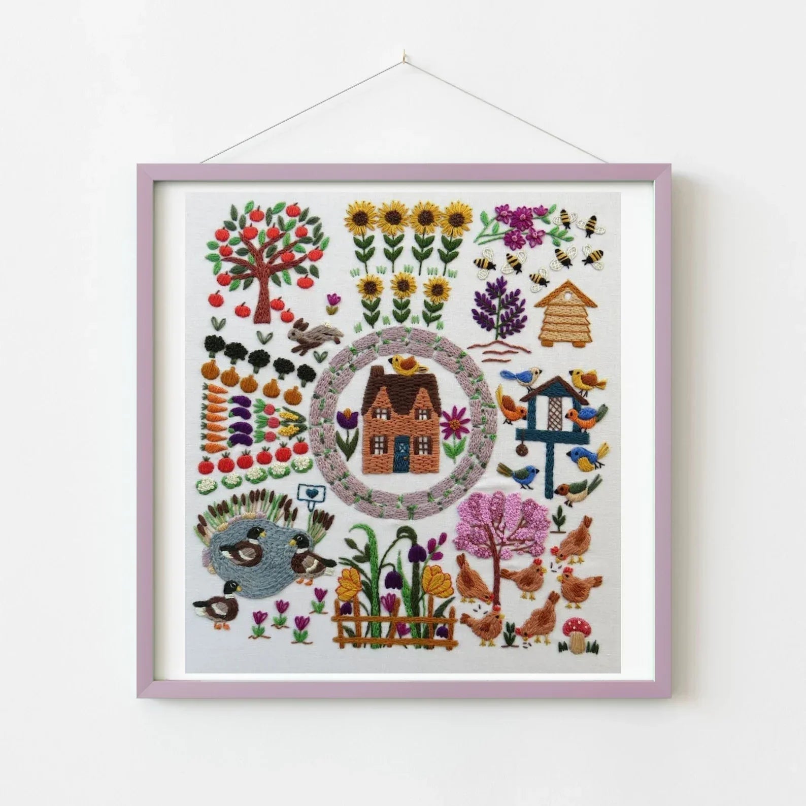 Embroidery of a garden scene with a house, flowers, and animals in a purple frame.