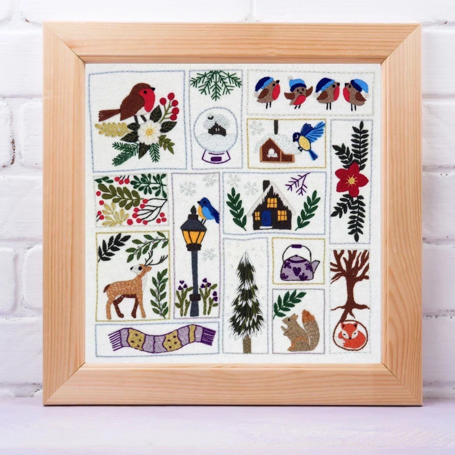 Framed cross-stitch pattern with nature-themed designs on a white brick wall background.