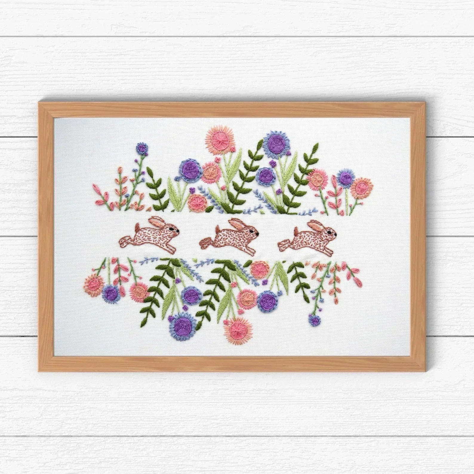 Embroidery design with rabbits and flowers on a white background with a wooden frame.