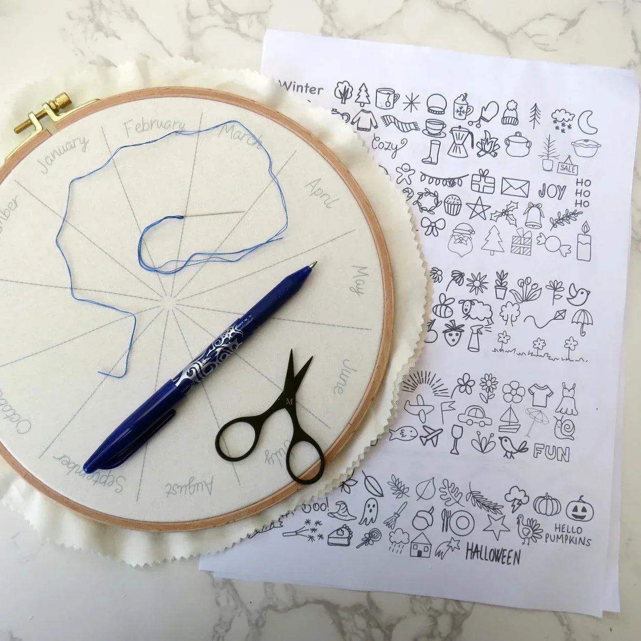 Embroidery hoop with pattern, pen, scissors, and a sheet of design ideas on a marble surface.