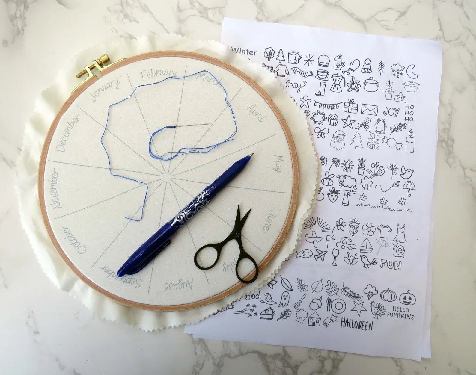 Embroidery hoop with pattern, pen, scissors, and a sheet of design ideas on a marble surface.