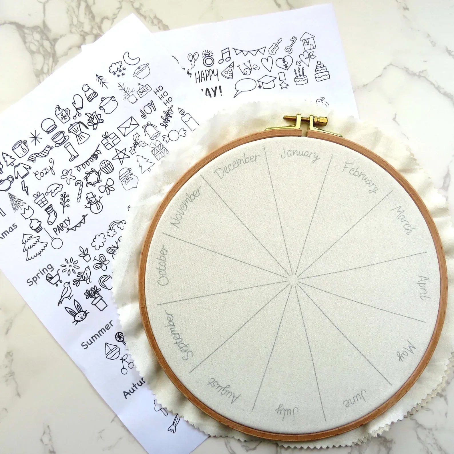 Embroidery hoop with a wheel of the year design on a marble surface with a sheet of doodles.