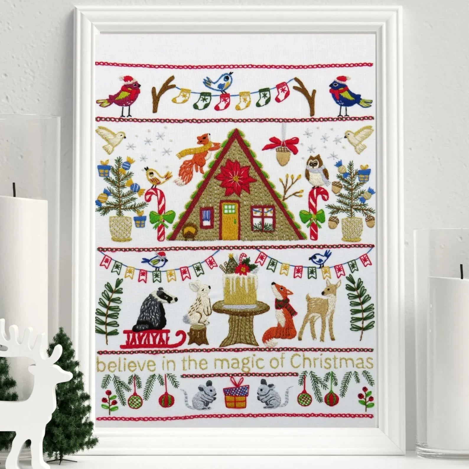 Framed cross-stitch art with Christmas-themed design featuring a cabin, animals, and festive elements.