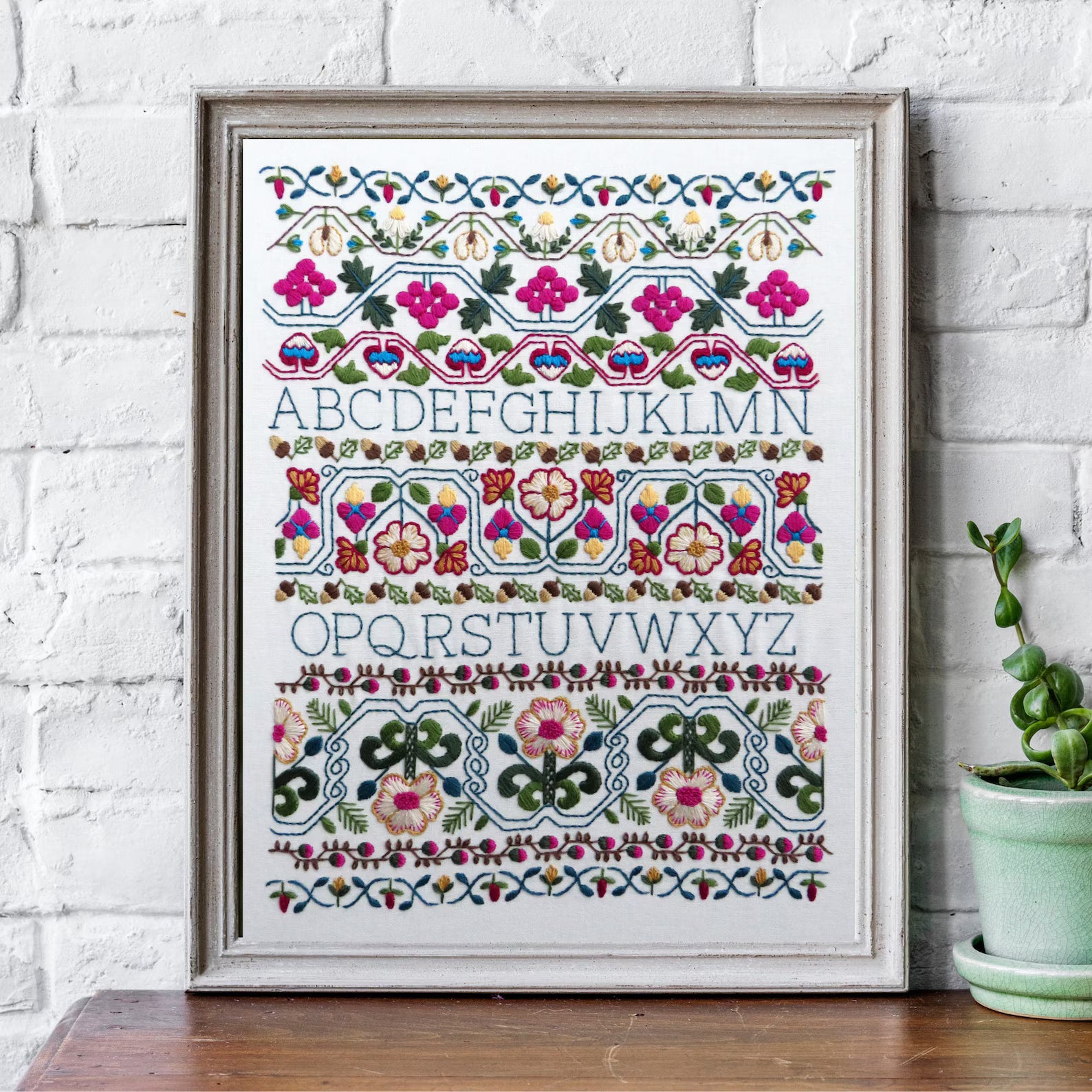 Framed cross-stitch alphabet chart with decorative elements on a white brick wall.