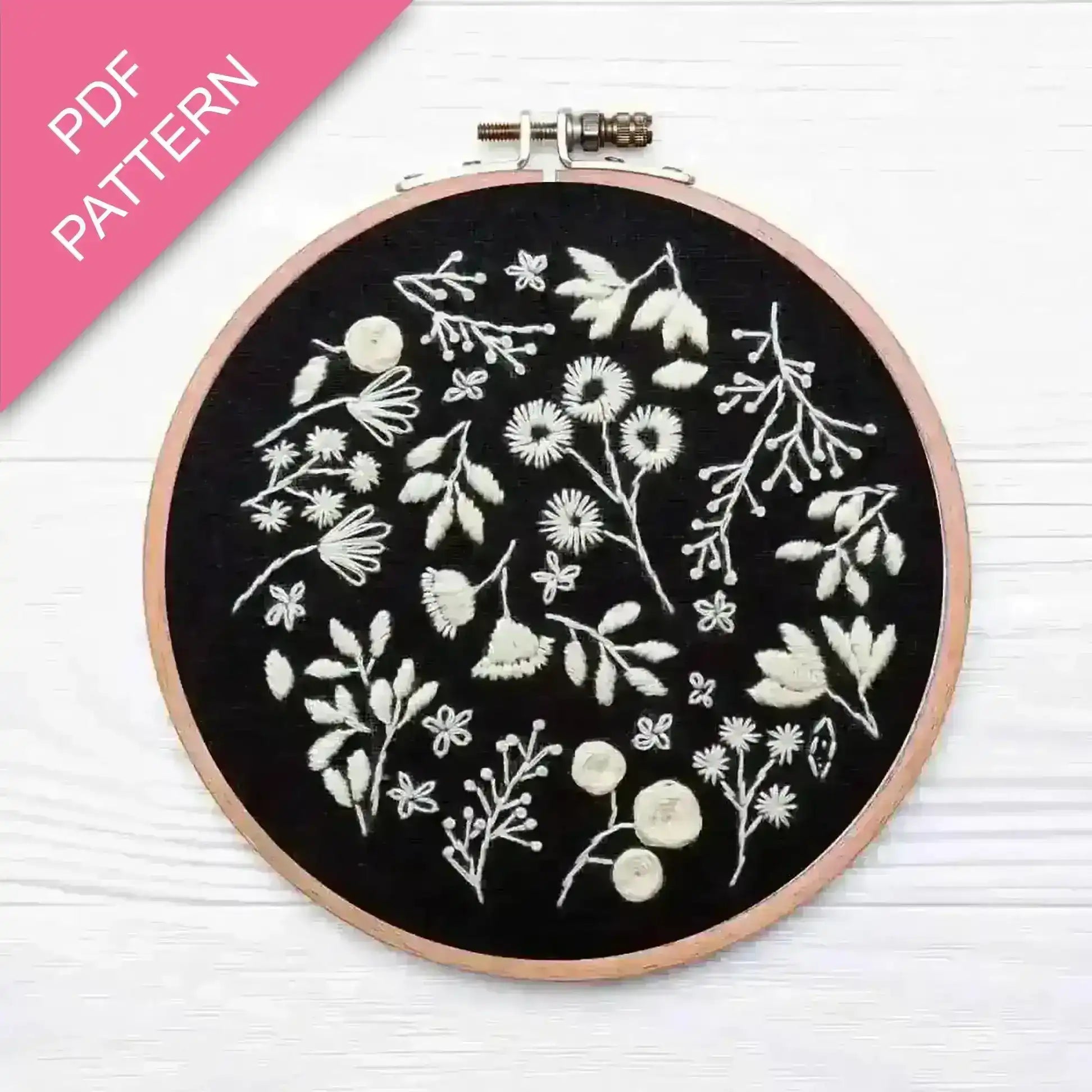 Embroidery hoop with white floral patterns on black fabric, pink PDF pattern label.