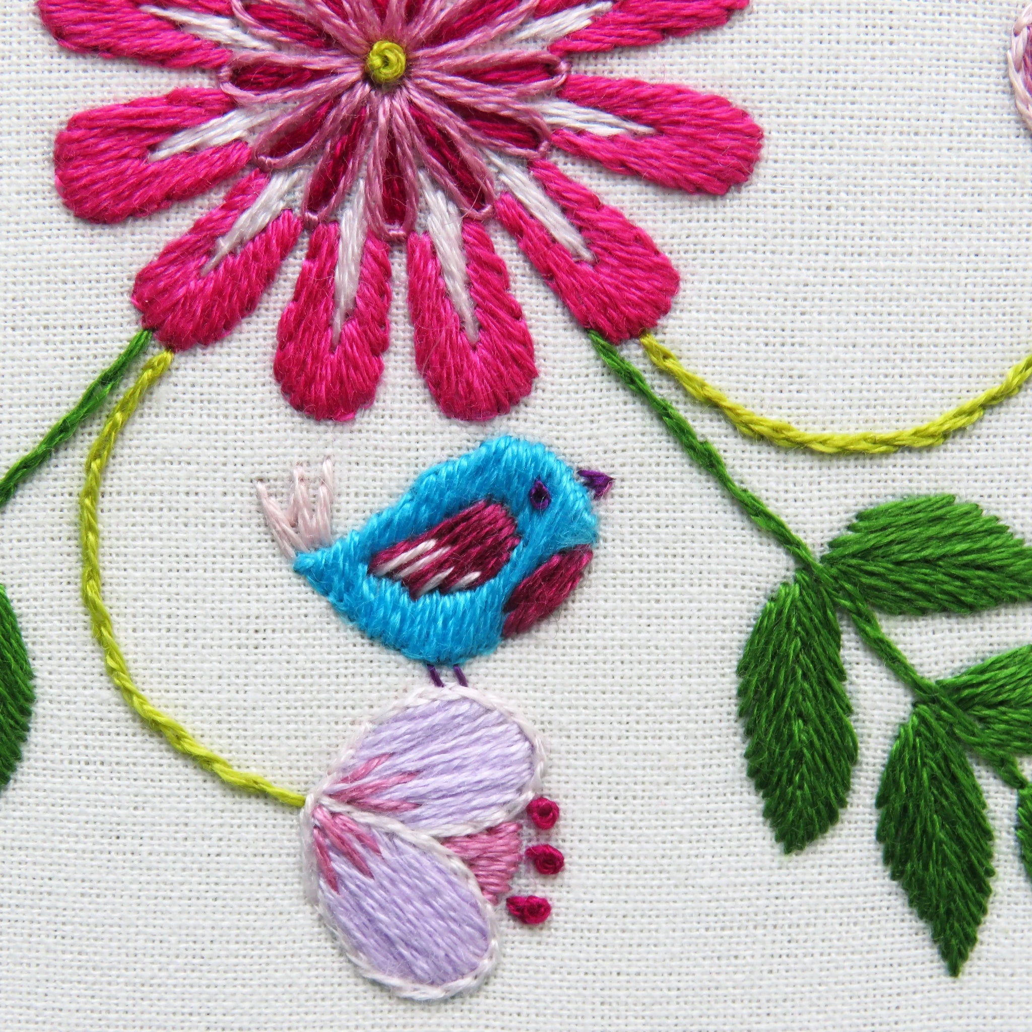 Embroidered design of a pink flower, blue bird, and green leaves on white fabric.