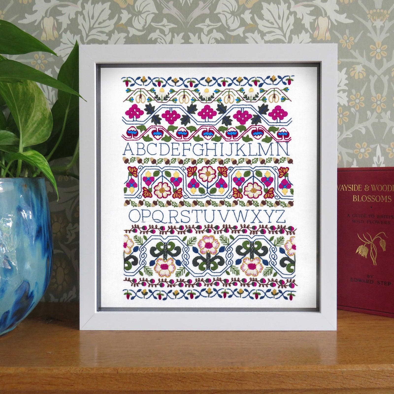Framed decorative alphabet print with floral patterns on a wooden surface.