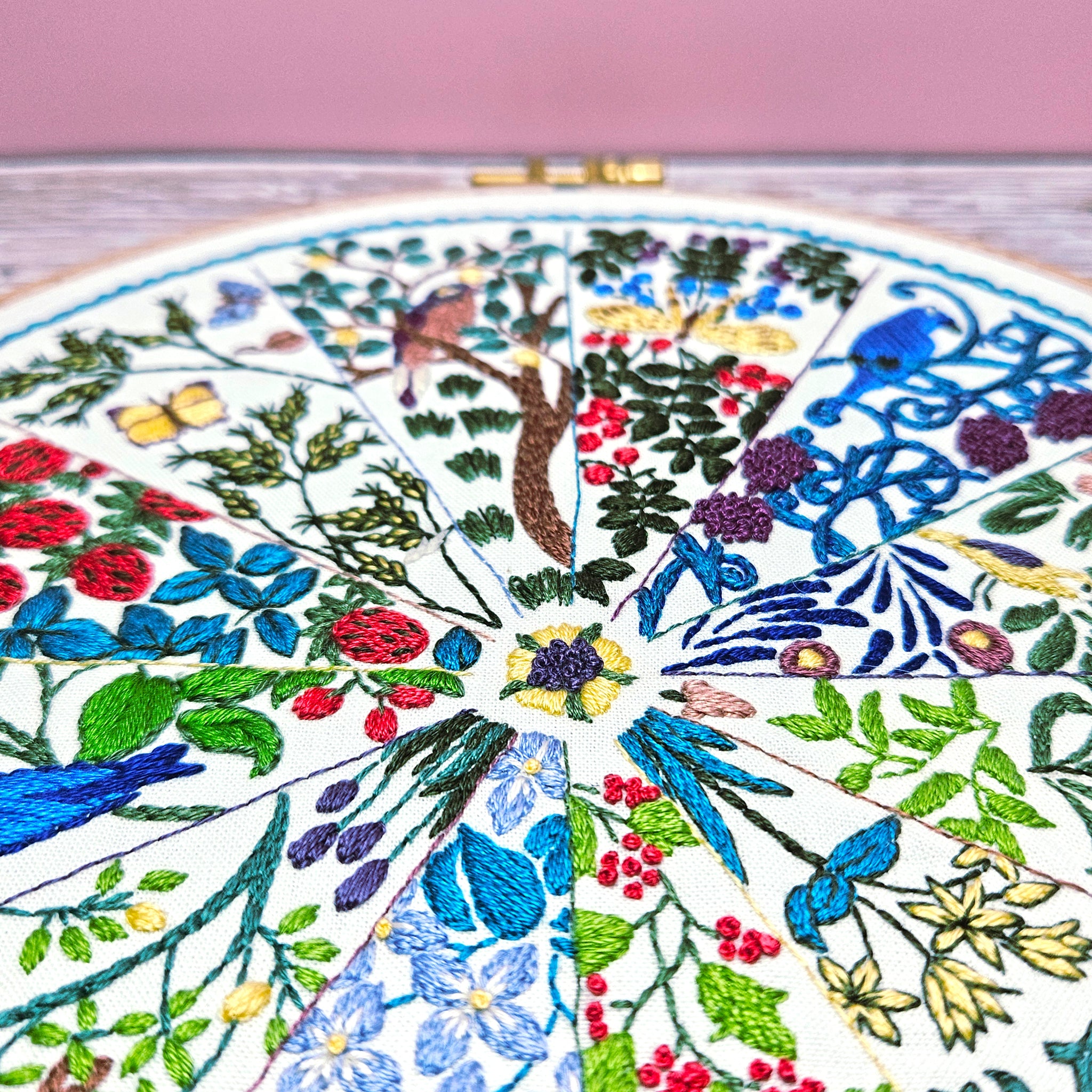 Embroidery hoop with colorful floral and nature-themed embroidery on a pink background