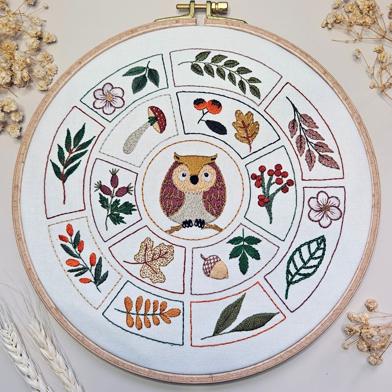 Embroidery hoop with owl design surrounded by leaves and flowers on a white background.
