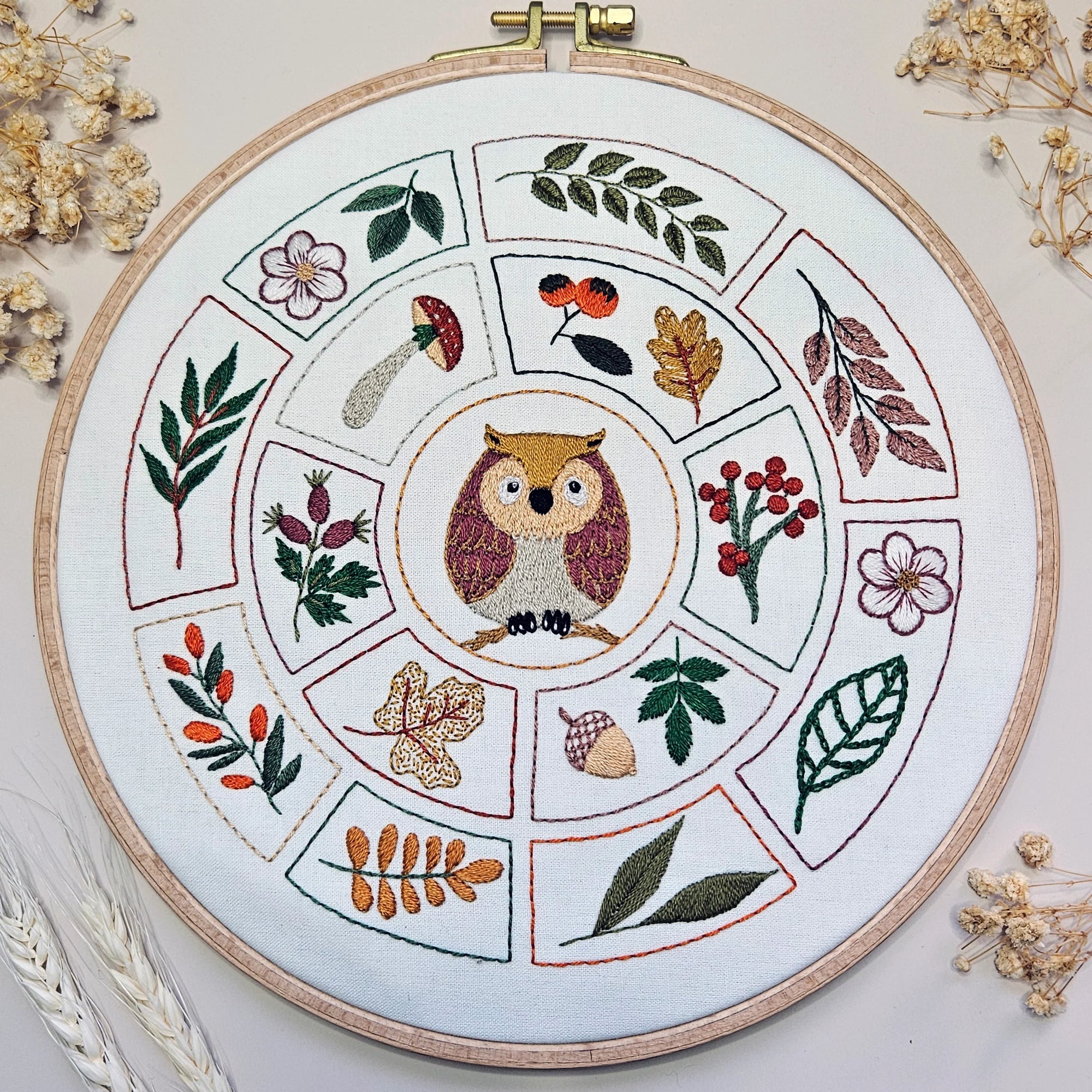 Embroidery hoop with owl design surrounded by leaves and flowers on a white background.