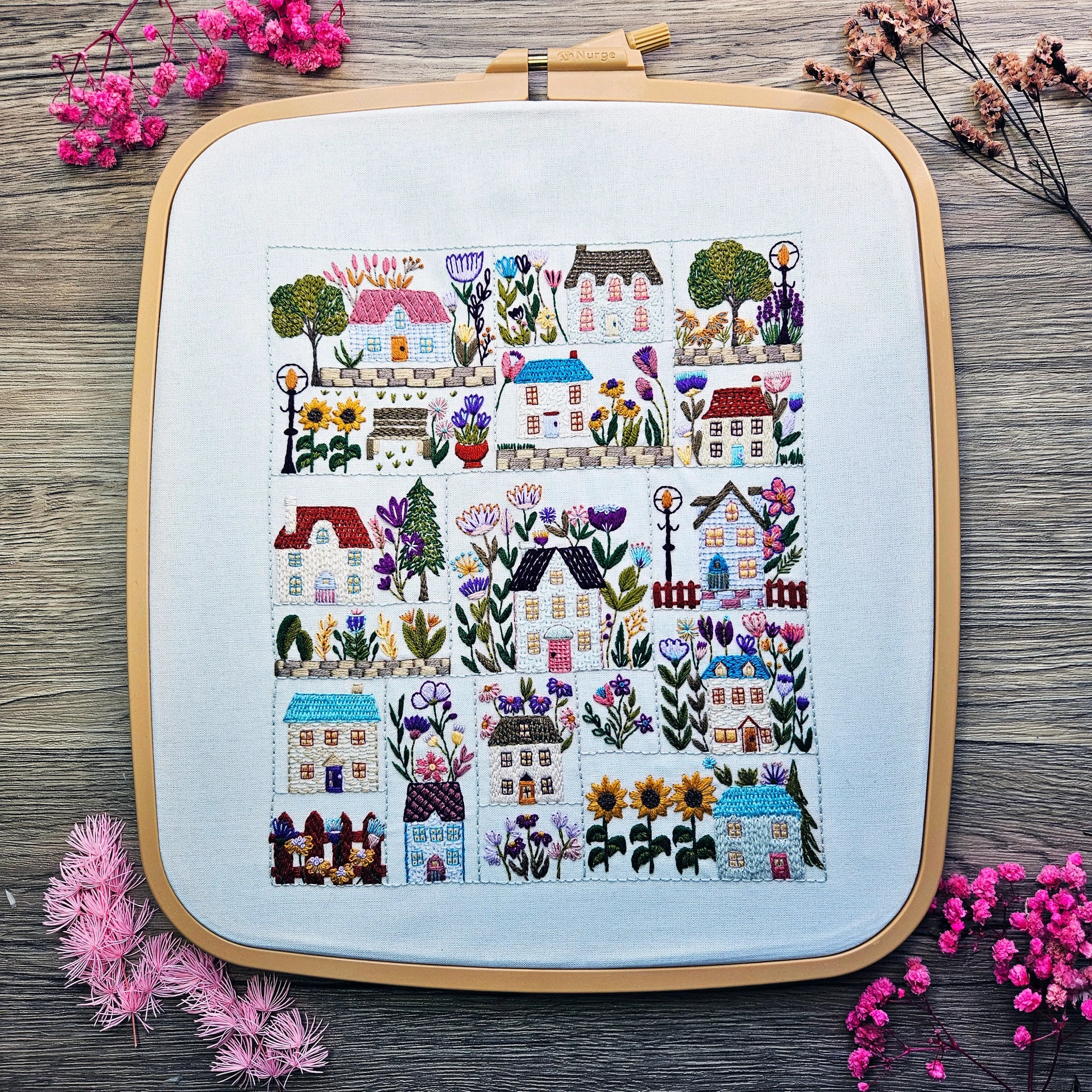 Embroidery hoop with a colorful stitched design of houses and flowers on a wooden surface with pink flowers.