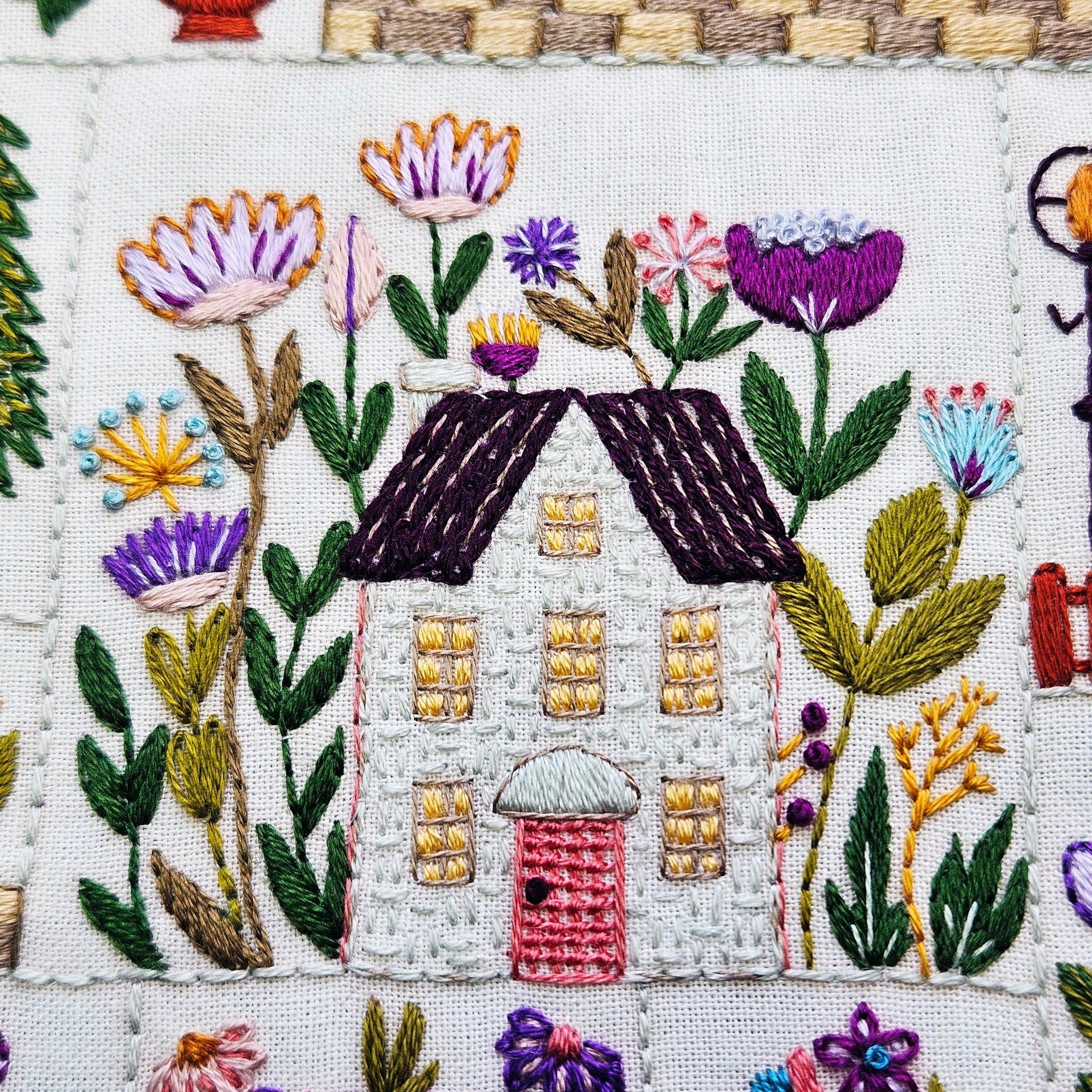 Embroidered design of a house surrounded by flowers and plants on a white background.