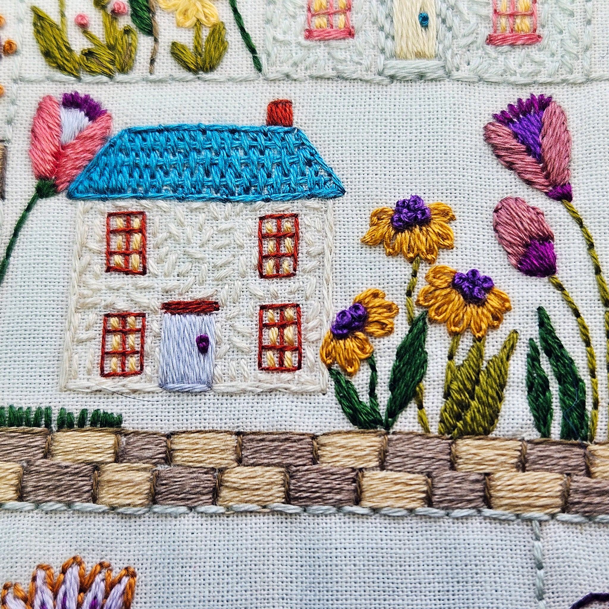 Embroidered scene with a house, flowers, and a fence on a white background