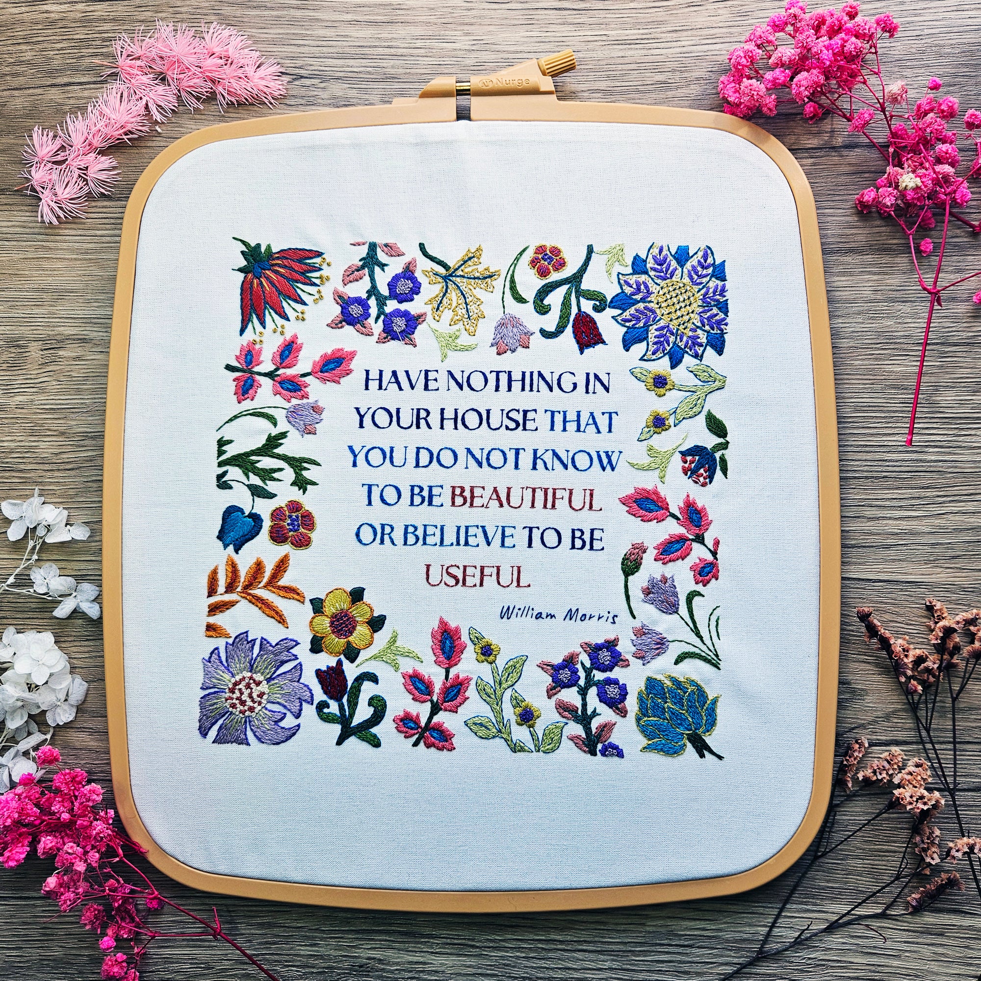 Embroidery hoop with floral design and text on a wooden surface with flowers.