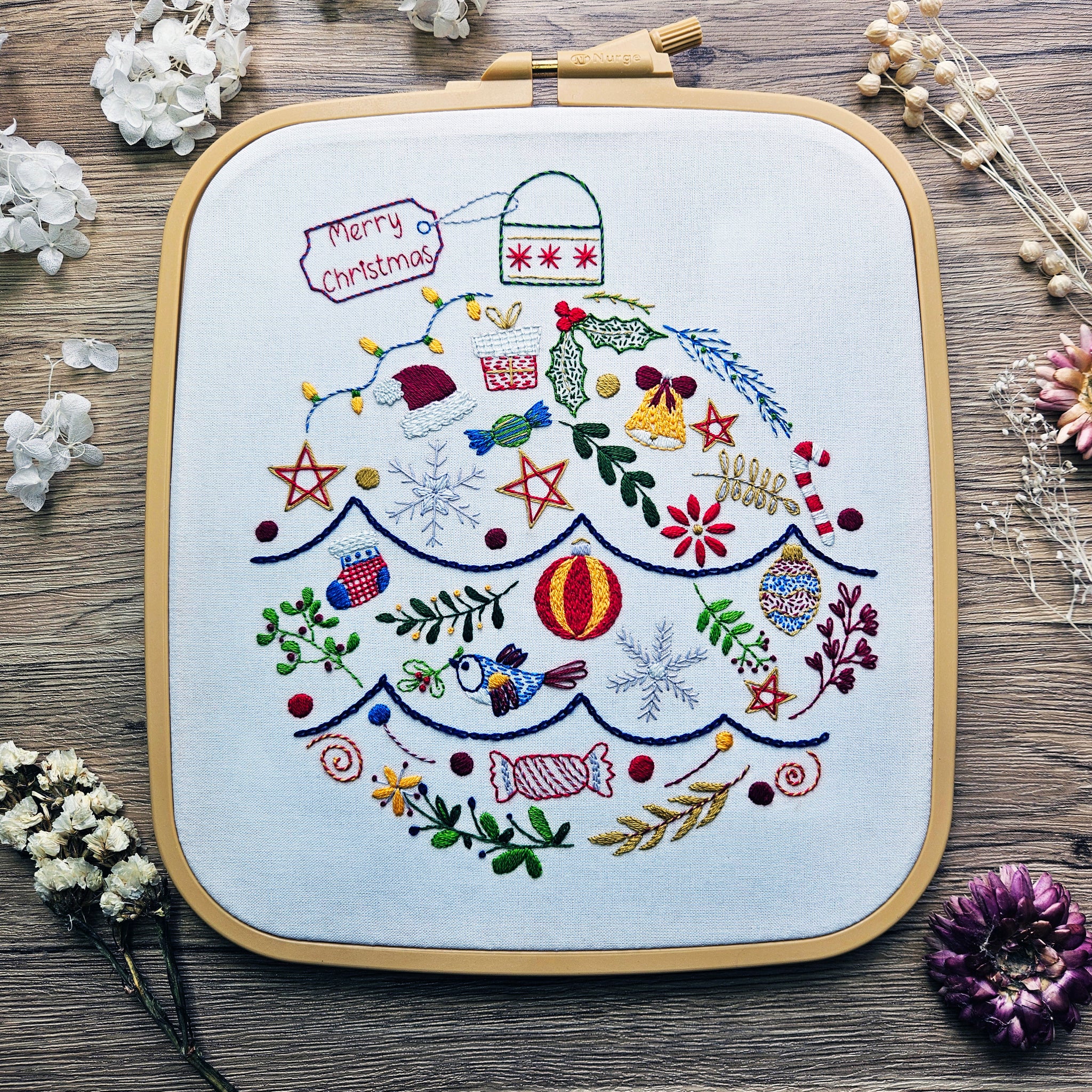 Embroidery hoop with a Christmas-themed embroidered design on a wooden surface with flowers.