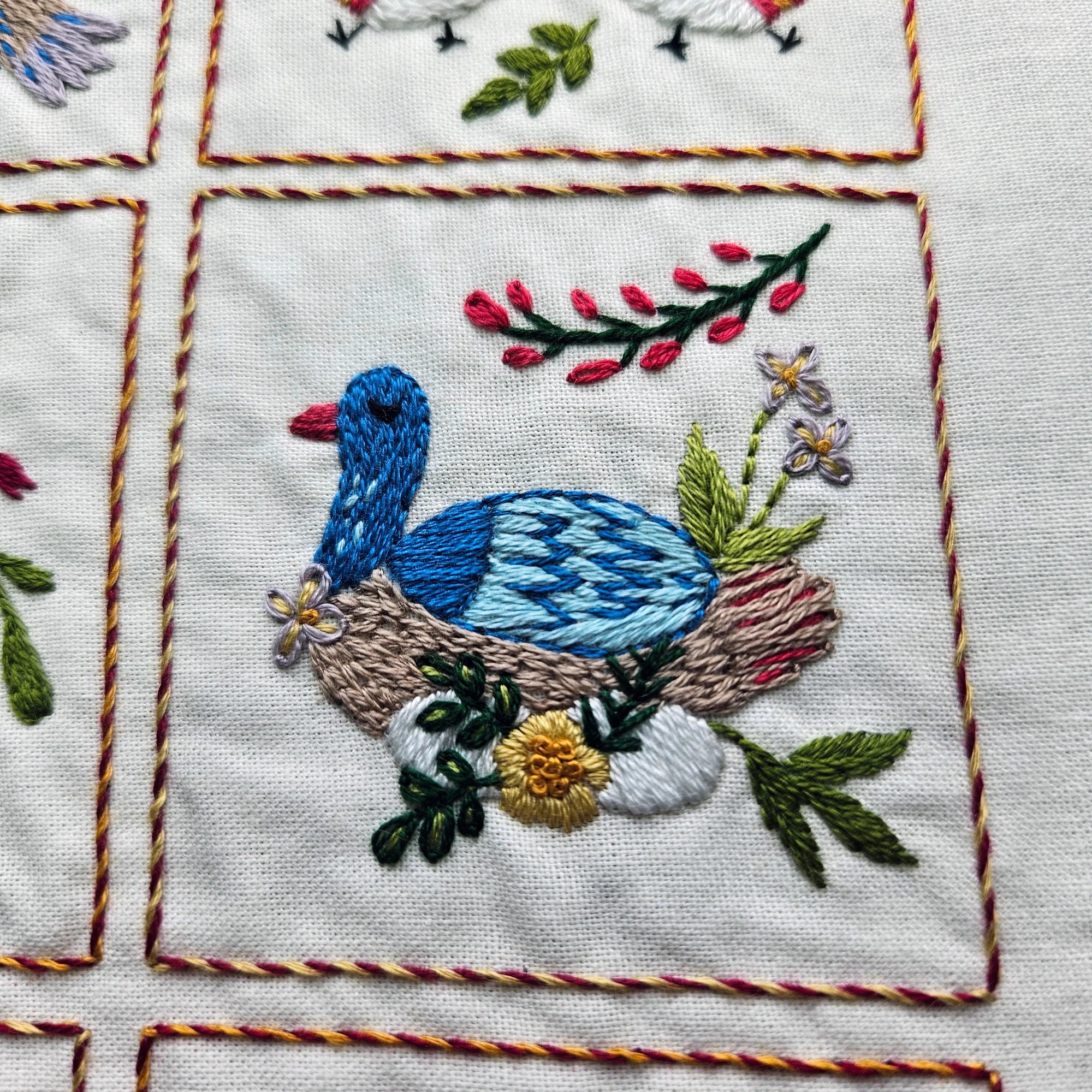 Embroidered design of a blue bird in a nest with flowers on a white fabric background.