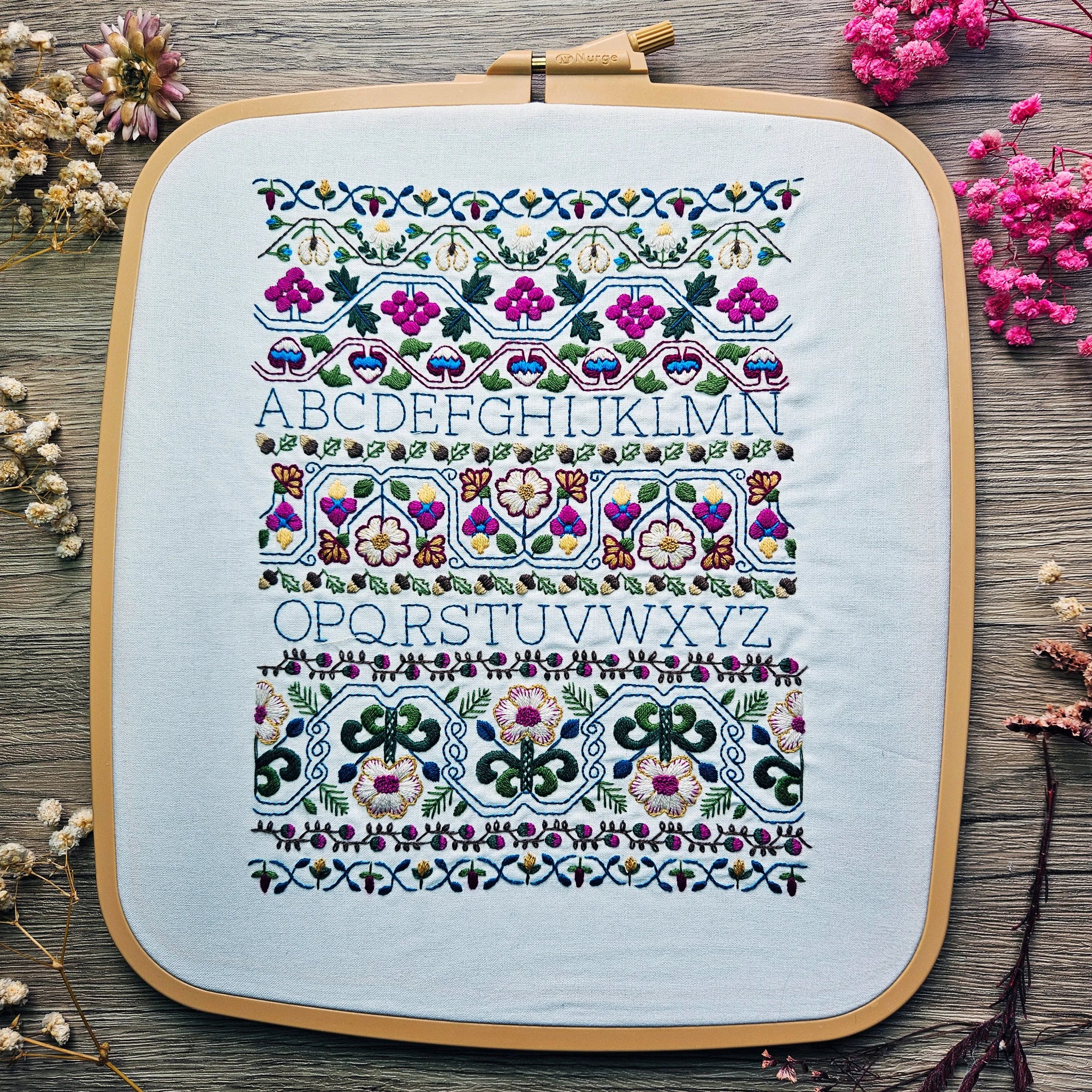 Embroidery hoop with floral alphabet pattern on a wooden surface with flowers.