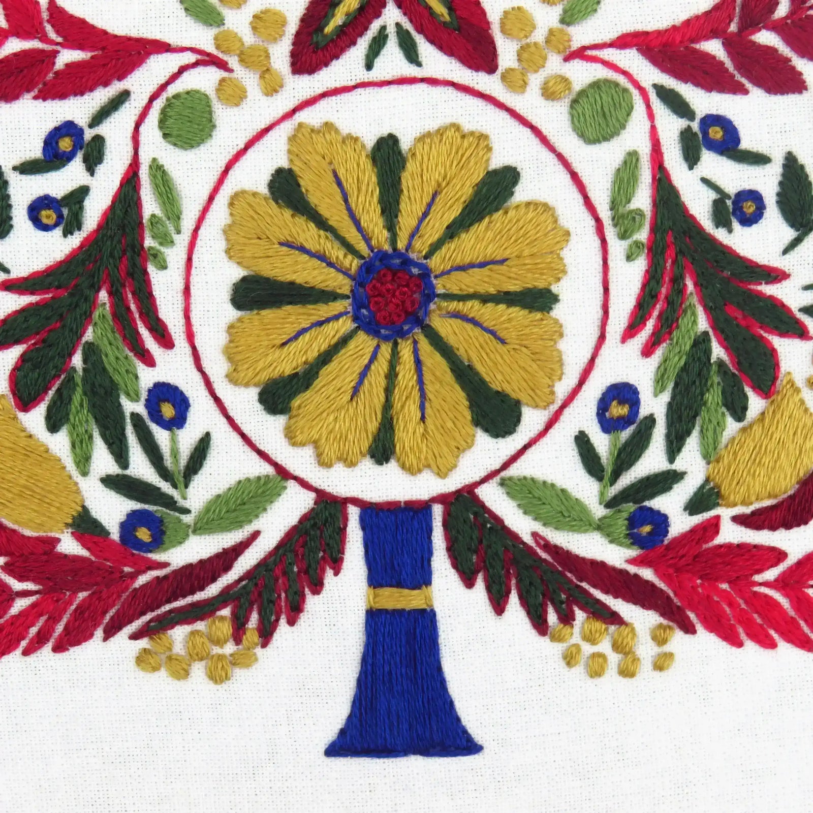 Close-up of an embroidered design with a central yellow flower surrounded by green leaves and red, blue, and gold accents on a white background.