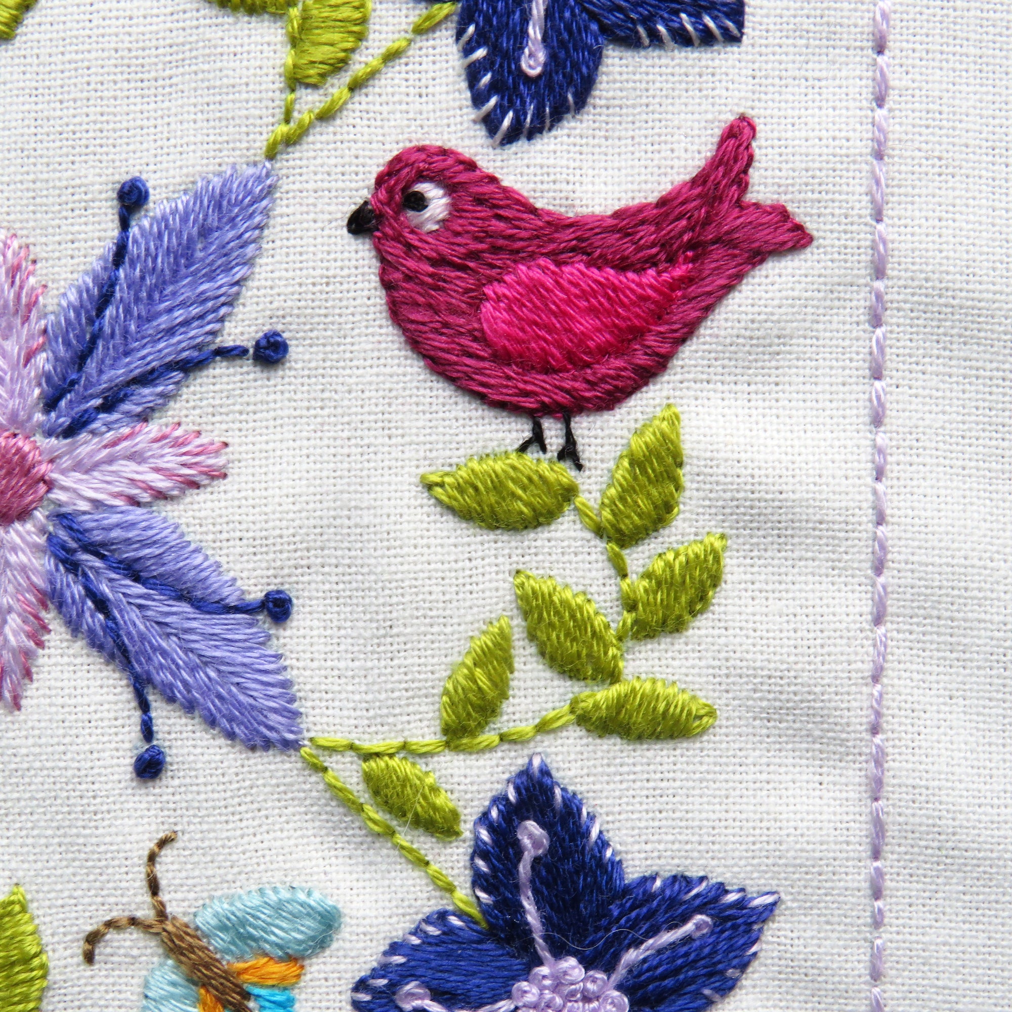Embroidered design with a pink bird, blue flowers, and green leaves on white fabric.