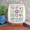 A framed floral embroidery sampler displayed on a small wooden stand on a wooden table, with a potted plant on the left and a book on the right, set against a patterned wallpaper background.