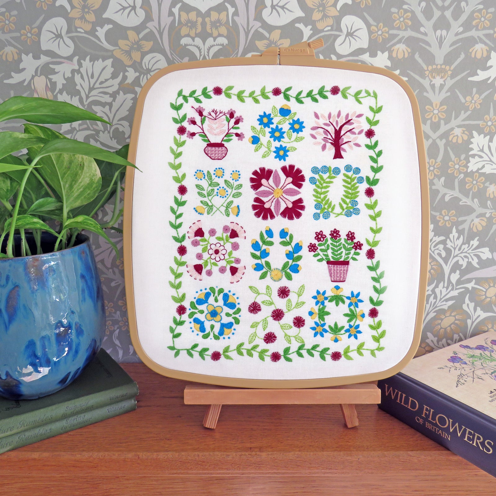 A framed floral embroidery sampler displayed on a small wooden stand on a wooden table, with a potted plant on the left and a book on the right, set against a patterned wallpaper background.