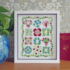 A framed floral cross-stitch or embroidery piece with a white mat and frame, displayed on a wooden surface against patterned wallpaper. A green plant in a blue vase is on the left, and a red book is on the right.