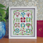 A framed floral cross-stitch or embroidery piece with a white mat and frame, displayed on a wooden surface against patterned wallpaper. A green plant in a blue vase is on the left, and a red book is on the right.