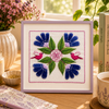 Framed cross-stitch embroidery of flowers and birds on a table with a notebook, pen, and cup.