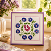 Decorative cross-stitch frame with flowers and a rooster on a table with a notebook, mug, and books.