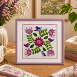 Framed cross-stitched floral design on a table with a mug, notebook, and candle.