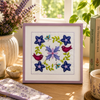 Framed cross-stitched artwork with flowers and birds on a table with books and a candle.