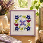 Framed cross-stitched artwork with flowers and birds on a table with books and a candle.