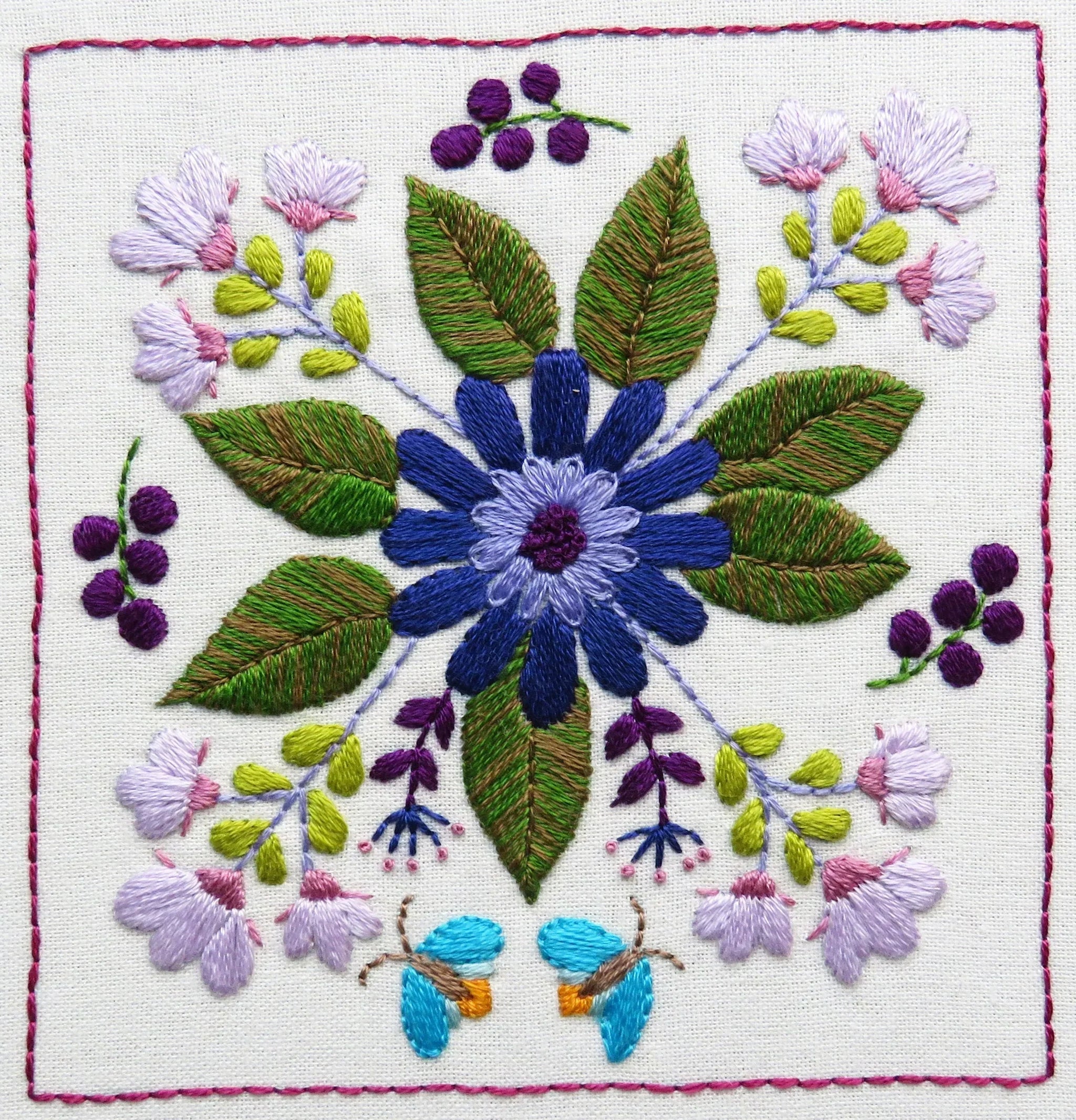 Embroidered design of a blue flower with green leaves and purple berries on a white background.