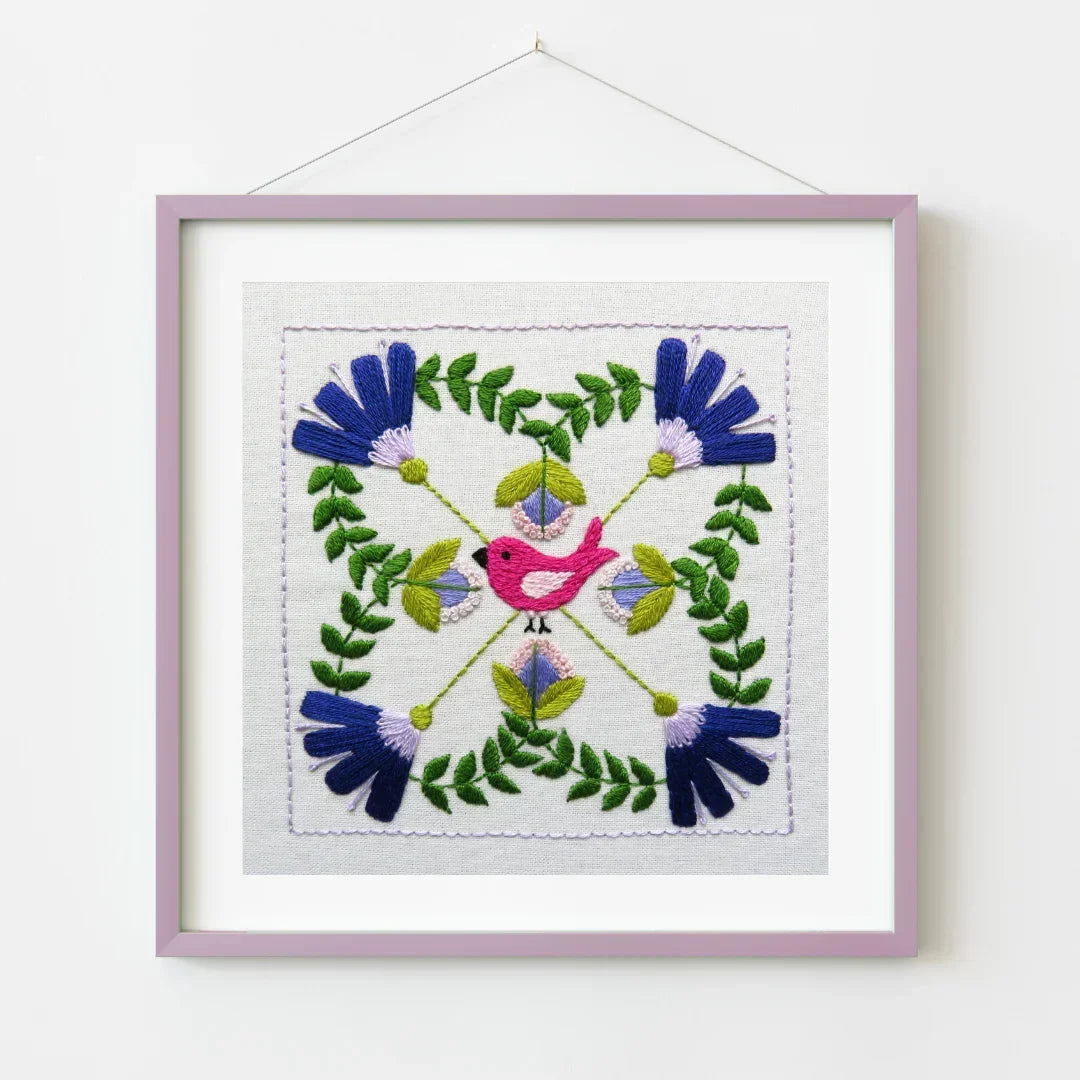 Embroidered square with a pink bird and floral patterns in a purple frame on a white background