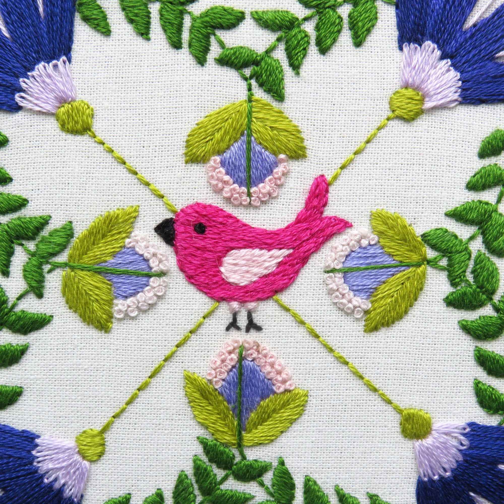Embroidered design with a pink bird surrounded by green leaves and purple flowers on a white background.