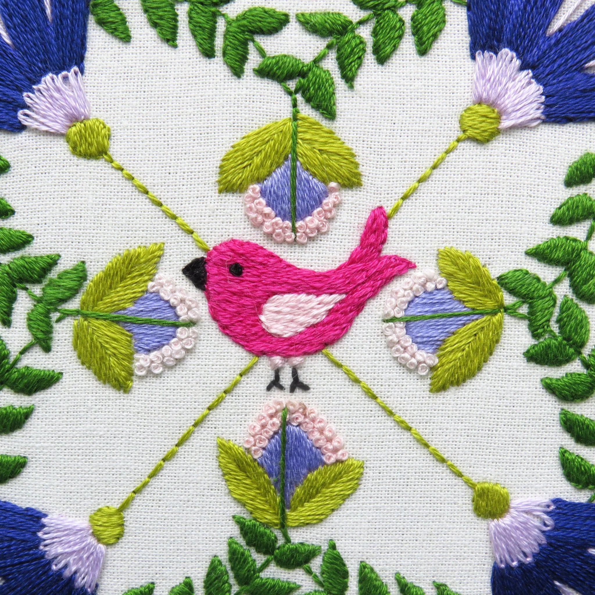 Embroidered design with a pink bird surrounded by green leaves and purple flowers on a white background.