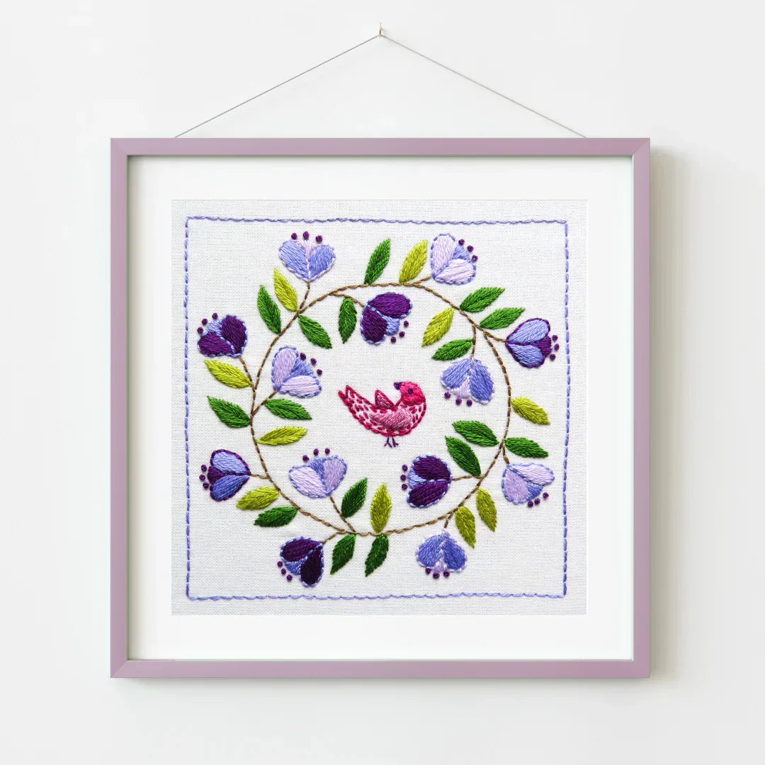 Embroidered artwork with a bird and floral design in a purple frame on a white background