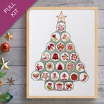 Framed cross-stitch pattern of a Christmas tree on a wooden surface with decorative items.