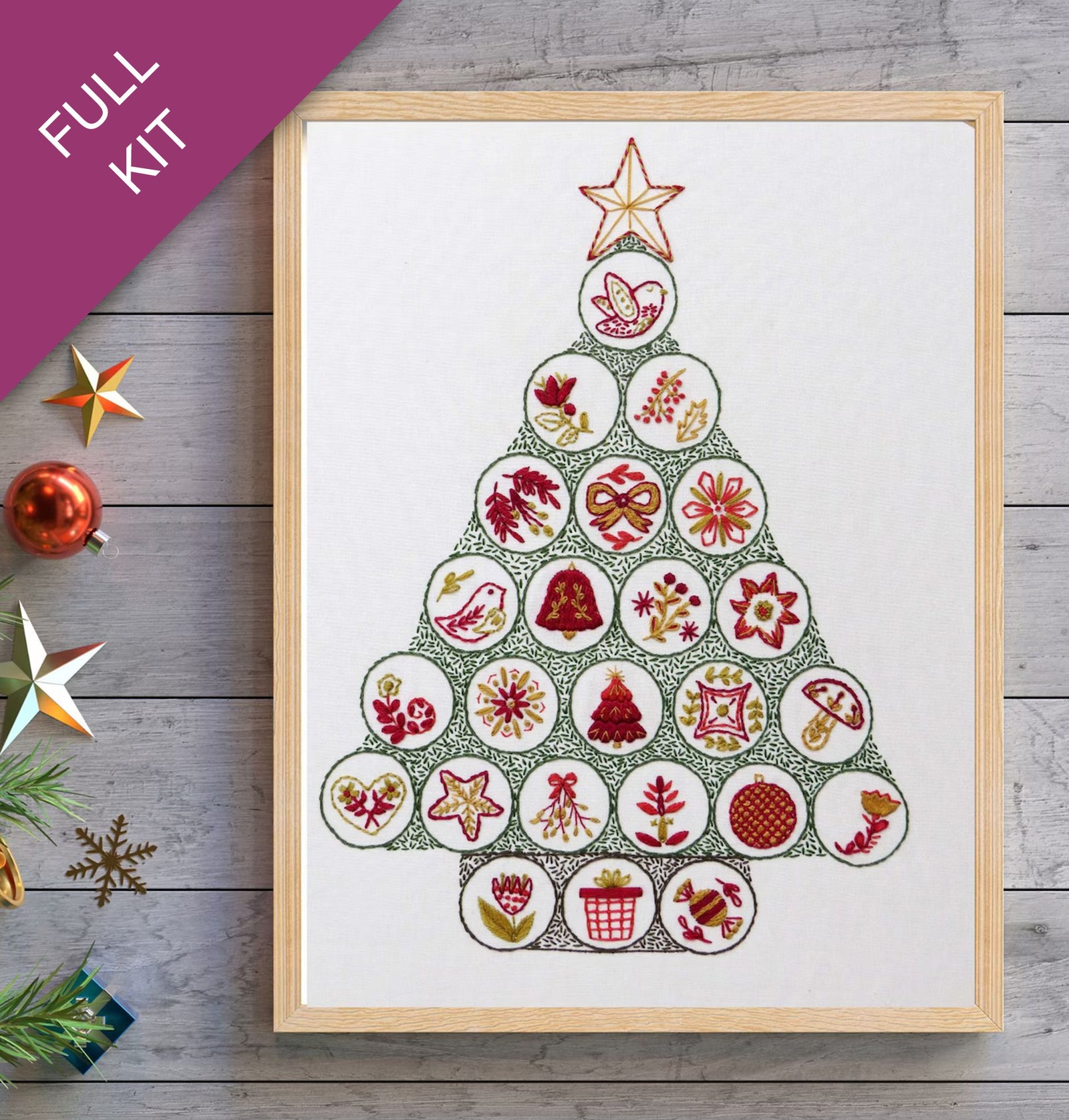 Framed cross-stitch pattern of a Christmas tree on a wooden surface with decorative items.