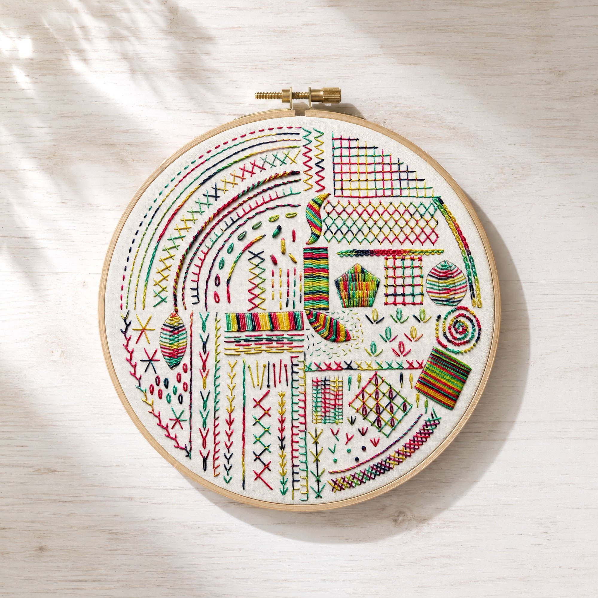 A round embroidery hoop holding a white fabric with colorful geometric and floral stitched patterns, photographed from above on a light wood surface with soft leaf shadows.