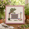 Embroidery of a garden scene with a tree, birds, and a checkered cloth, framed in pink.